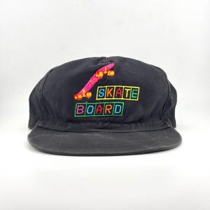 May include: A black baseball cap featuring an embroidered graphic of a pink skateboard above the text SKATE BOARD. The word SKATE is in yellow letters within green boxes, and the word BOARD is in multi-coloured letters within green boxes.