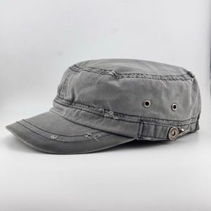 Puede incluir: Una gorra de estilo militar gris carbón con parte superior plana, visera corta y curva, y bordes desgastados y deshilachados. La gorra incluye dos ojales de ventilación de metal plateado en el lateral y un pequeño detalle de botón metálico cerca de la visera.