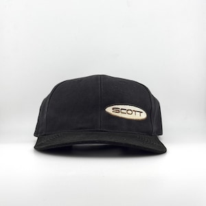 Puede incluir: Gorra de béisbol negra con visera curva. La gorra presenta un parche ovalado bordado con el nombre de la marca "SCOTT" en letras blancas sobre un fondo beige. La gorra está sobre un fondo blanco.