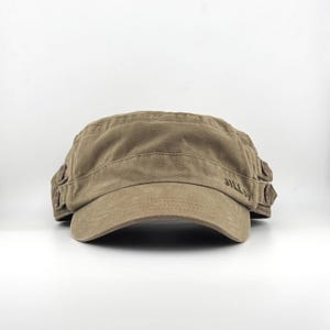 May include: A tan military-style cap with a curved brim. The cap has a structured crown with stitched details and a small, curved visor. The text "JILL by" is visible on the side. The cap is made of a soft, durable fabric.