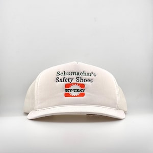 May include: White trucker hat with the text "Schumacher's Safety Shoes" in green and a red and orange logo that says "HY-TEST". The hat has a white mesh back and a curved brim.