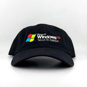 May include: A black baseball cap featuring an embroidered logo on the front. The logo includes the colourful four-pane window icon and the white text Microsoft Windows XP Tablet PC Edition with the letters XP in red.