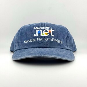 May include: A blue denim baseball cap featuring the embroidered white text Microsoft .net and Services Platform Division. The logo includes small blue, green, yellow, and red square accents above the letters n, e, and t on the front panel.