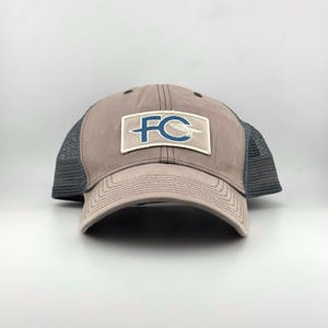 Op de afbeelding: Een taupekleurige baseballpet met een donkergrijze mesh achterkant en een rechthoekige patch op de voorkant. De patch bevat de blauwe letters FC met een gestileerde witte vogelafbeelding in het ontwerp, op een lichtcrème achtergrond.