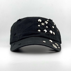 Puede incluir: Una gorra de estilo militar negra con detalles de tela desgastada y un surtido de tachuelas metálicas plateadas, incluyendo formas redondas, cuadradas y de estrella. La visera está adornada con ojales y tachuelas metálicas adicionales.