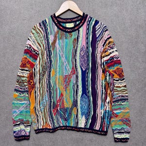 May include: A colorful, patterned sweater with a crew neck and long sleeves. The sweater features a variety of vertical stripes in shades of blue, green, red, orange, and purple. The sweater is hanging on a wooden hanger.