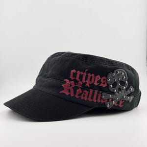 以下が含まれることがあります： 側面に赤い文字で「cripes Reallize」とプリントされた黒いミリタリースタイルのキャップ。小さな銀色の金属製スタッドで装飾された灰色のスカルとクロスボーンのアップリケが、右側のテキストの上に配置されています。