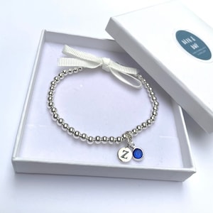 May include: A silver beaded bracelet with a charm featuring the letter "Z" and a blue gemstone. The bracelet is in a white gift box with a white ribbon.