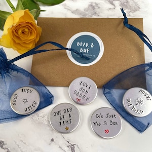 May include: Five silver-toned metal tokens with inspirational messages, including "Good Luck Aster", "Love You Daddy", "One Day At A Time", "It's Just Me & You", and "You've Got This!" The tokens are displayed on a blue and brown background.