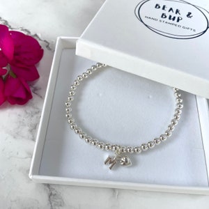 May include: A silver bracelet with a heart charm and a letter 'M' charm. The bracelet is in a white box with the text 'BEAR & BUP HAND STAMPED GIFTS'.