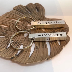 May include: Two silver keyrings with rectangular bars. One reads "Our first kiss" and the other displays coordinates and a heart symbol. The keyrings are on a bronze-coloured leaf-shaped dish.