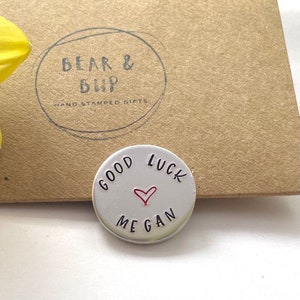 May include: A circular, silver-toned metal token with the words "GOOD LUCK MEGAN" stamped on it, and a small red heart. The token is in front of a brown cardboard box with the text "BEAR & BUP HAND STAMPED GIFTS" printed on it.