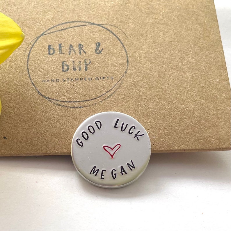 May include: A circular, silver-toned metal token with the words "GOOD LUCK MEGAN" stamped on it, and a small red heart. The token is in front of a brown cardboard box with the text "BEAR & BUP HAND STAMPED GIFTS" printed on it.