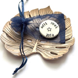 May include: A round silver-toned metal token stamped with the text WELL DONE and JOSH separated by a star symbol. The token rests on a dark blue sheer organza drawstring pouch, which is placed atop a gold-colored metal leaf-shaped decorative tray.