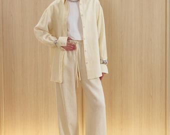May include: A cream-colored linen outfit consisting of a long-sleeved button-down shirt, a white t-shirt, and wide-leg pants. The outfit is paired with sandals and silver jewelry. The background features light wood paneling and a wooden floor.