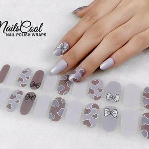 May include: Nail polish wraps with a light gray base and a design of white hearts and bows. The wraps are shown on a model's fingernails. The text "NailsCool" and "Nail Polish Wraps" is visible in the image.