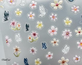 May include: A sheet of nail art stickers featuring a variety of white, pink, and yellow flowers with green stems and leaves, and white, blue, and black butterflies. The stickers are on a clear background.