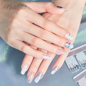 May include: A hand model with white and silver nail polish designs. The nails are long and oval shaped. The designs include glitter, stars, and abstract patterns. The image also shows a sheet of nail polish stickers with a similar design.