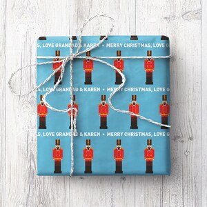May include: A wrapped gift with light blue paper decorated with red toy soldier illustrations. The gift is tied with white string and has the text "MERRY CHRISTMAS, LOVE G".