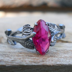 Puede incluir: Anillo de plata con una llamativa gema rosa brillante con corte marquesa. La banda del anillo presenta detalles de hojas y vides, creando un diseño inspirado en la naturaleza. La gema es el foco principal, engastada con garras.