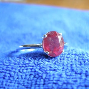 Puede incluir: Un anillo de plata con una gran piedra preciosa de rubí de forma ovalada. El rubí es de color rojo intenso y tiene un corte facetado.