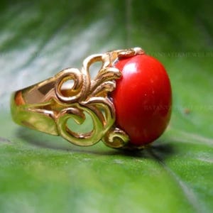 May include: A gold-colored ring featuring an oval, bright red gemstone. The ring has an ornate, openwork design on the sides, with a polished finish. The gemstone is the focal point, set against a green background.