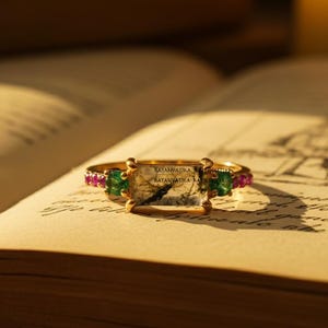 May include: A gold ring with a rectangular central stone bearing the text "RATANVATIKA". The ring is adorned with green and pink gemstones. The ring is displayed on the open pages of a book.