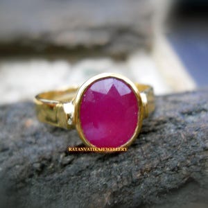 May include: A gold ring with a large, oval-cut ruby gemstone. The ruby is a deep pink color and is set in a gold bezel. The ring has a simple, classic design. The text "RATANVATIKAJEEWELLERY" is visible on the ring.