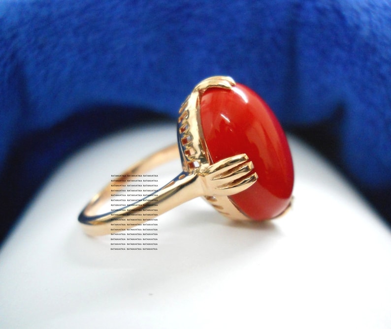 May include: A gold ring with a large, oval, bright red gemstone. The ring has a gold-coloured band and setting, with intricate detailing around the gemstone. The ring is set against a blue background.