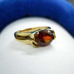 May include: A gold ring with a large, oval-cut, reddish-brown gemstone. The ring's band is a polished gold, and the gemstone is set in a decorative gold setting. The ring is displayed on a white surface with a blue background.
