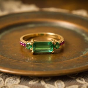 May include: A gold ring with a rectangular green emerald center stone and two smaller green emeralds on either side. The ring also features small pink gemstones. The ring is displayed on a bronze-colored plate.