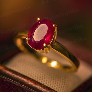 May include: A gold ring featuring a large, oval-cut ruby gemstone. The ruby is a deep red color and is set in a four-prong setting. The ring's band is a simple, polished gold.