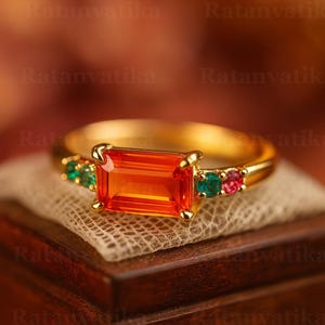 May include: A gold ring featuring a large, rectangular orange gemstone as the centerpiece. The ring is accented with smaller green and pink gemstones on either side of the main stone. The ring is displayed on a wooden surface.