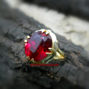 Könnte beinhalten: Ein goldener Ring mit einem großen, oval geschliffenen, tiefroten Edelstein. Der Edelstein ist in einer goldenen Lünette mit dekorativen Zinken gefasst. Der Ring wird auf einer dunklen, strukturierten Oberfläche präsentiert. Der Text "RATANVATIKAJEWELLERY" ist sichtbar.