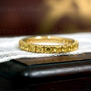 Könnte beinhalten: Ein goldener Ring mit einer durchgehenden Reihe gelber Edelsteine. Der Ring liegt auf weißer Spitze, mit einer dunklen Holzoberfläche im Hintergrund. Der Ring ist ein Voll-Eternity-Ring.