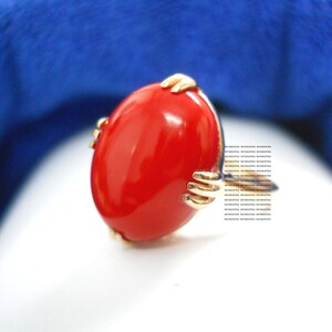 May include: A gold ring with a large, oval, bright red stone. The gold setting has a unique design, with small, hand-like structures holding the stone. The ring is set against a white and blue background.
