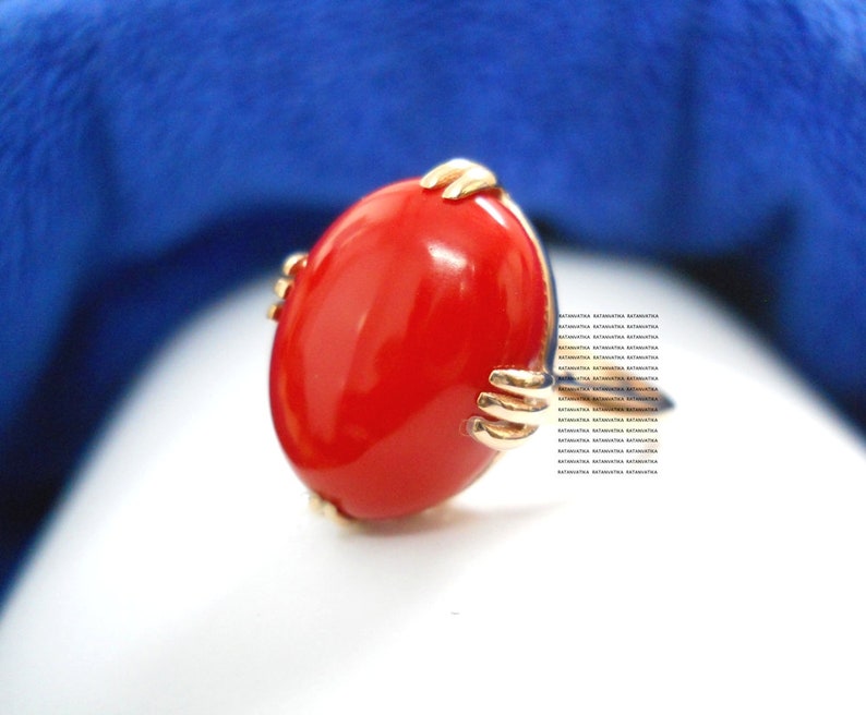 May include: A gold ring with a large, oval, bright red stone. The gold setting has a unique design, with small, hand-like structures holding the stone. The ring is set against a white and blue background.