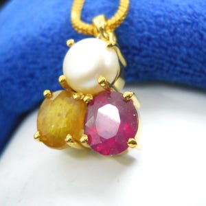 May include: A gold pendant necklace featuring a pearl and two gemstones. The gemstones are a ruby and a yellow sapphire. The pendant is set in gold and hangs from a gold chain. The background is a blue fabric.
