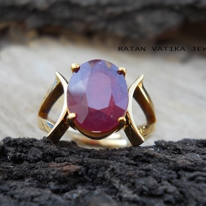 Könnte beinhalten: Ein goldener Ring mit einem großen, ovalen Rubin-Edelstein. Der Ring ist mit einem schlichten, eleganten Design besetzt.  RATAN VATIKA JEWELRY ist im Hintergrund gedruckt.