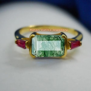 Puede incluir: Un anillo de oro con una piedra preciosa de ágata verde musgo y dos piedras de acento de rubí rojo. El anillo está engastado en una banda de oro y tiene un acabado pulido. El texto 'Ratan Vatika Jewellery' está impreso en el anillo.
