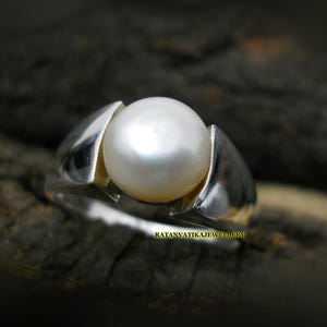 Puede incluir: Anillo de plata con una gran perla blanca y brillante. La perla se encuentra entre dos bandas plateadas curvas. El anillo se muestra sobre un fondo oscuro y texturizado, resaltando la elegancia de la joya.