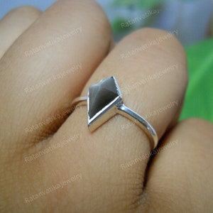 May include: A silver ring with a dark gray, faceted gemstone set in a geometric shape. The ring is on a person's finger.