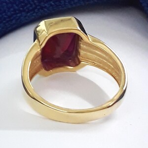 May include: A gold ring with a large, dark red, rectangular gemstone. The ring has a wide band with a textured design.