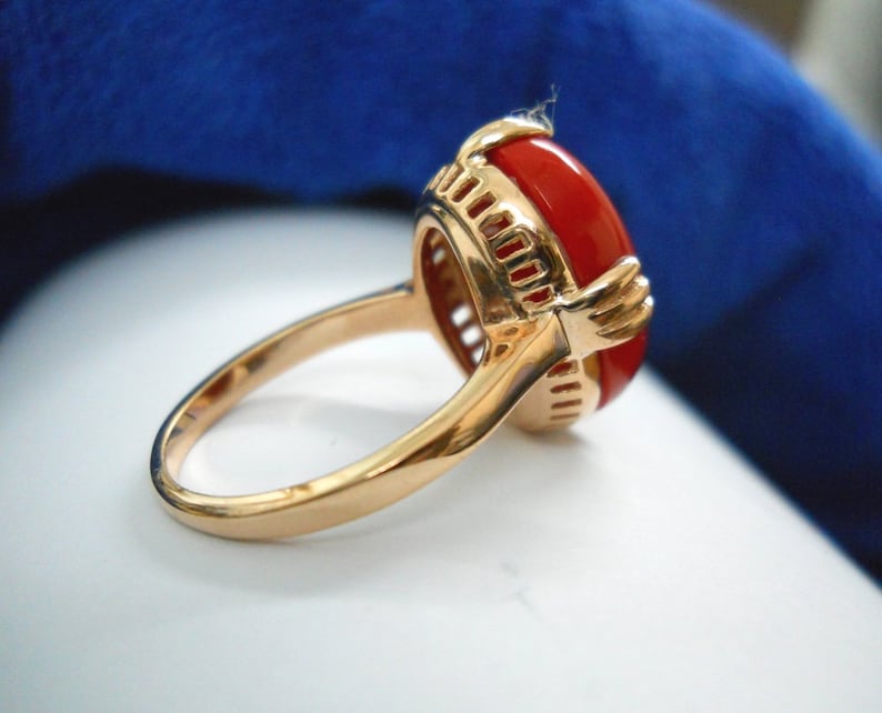 May include: Gold ring with a large, oval, red gemstone. The ring has a detailed gold setting with an openwork design around the gemstone. The band is smooth and polished, with a classic, elegant style.