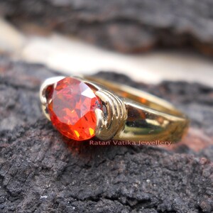 May include: A gold ring featuring a large, oval-cut, fiery orange gemstone. The ring's band is a polished gold, and the gemstone is set in a secure, decorative bezel. The text "Ratan Vatika Jewellery" is visible.