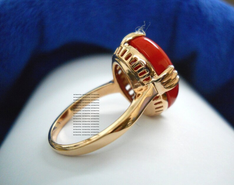 May include: A gold ring featuring a large, oval, red gemstone. The ring has an intricate, openwork design surrounding the gemstone and a polished band. The gemstone is a bright, striking red colour.