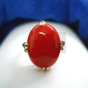 May include: A gold ring with a large, oval, glossy red stone. The ring has small gold accents and small blue stones. The background is a gradient of white and blue.