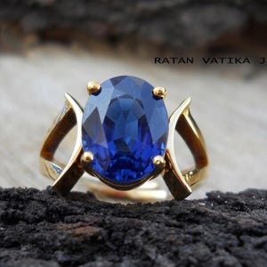 May include: A gold ring featuring a large, oval-cut, deep blue gemstone. The ring has a unique design with curved gold accents on either side of the stone. The ring is set against a dark, textured background. The text "RATAN VATIKA JEWEL" is visible.