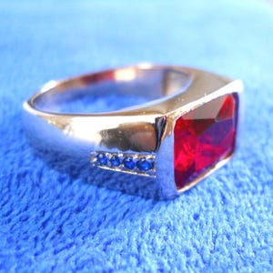 May include: A silver ring featuring a large, rectangular, red gemstone and a row of smaller, blue gemstones.