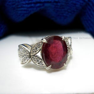 Può includere: Un anello d'argento con una grande pietra preziosa ovale di colore rosso scuro circondata da piccole pietre preziose chiare. L'anello ha un design floreale delicato.